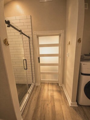 Un couloir avec une douche en verre, une porte blanche à panneaux dépolis et un lave-linge.