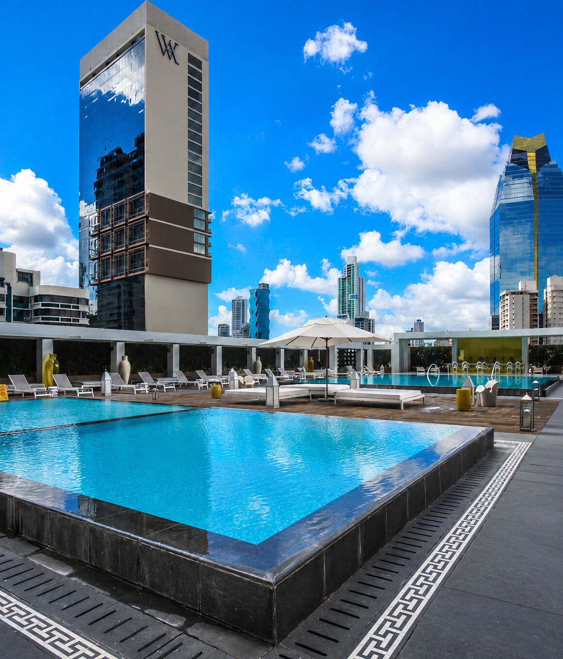 A rooftop pool with lounge chairs, a modern building with a reflective facade, and a clear blue sky.