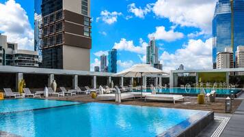 A rooftop pool with lounge chairs, a modern building with a reflective facade, and a clear blue sky.