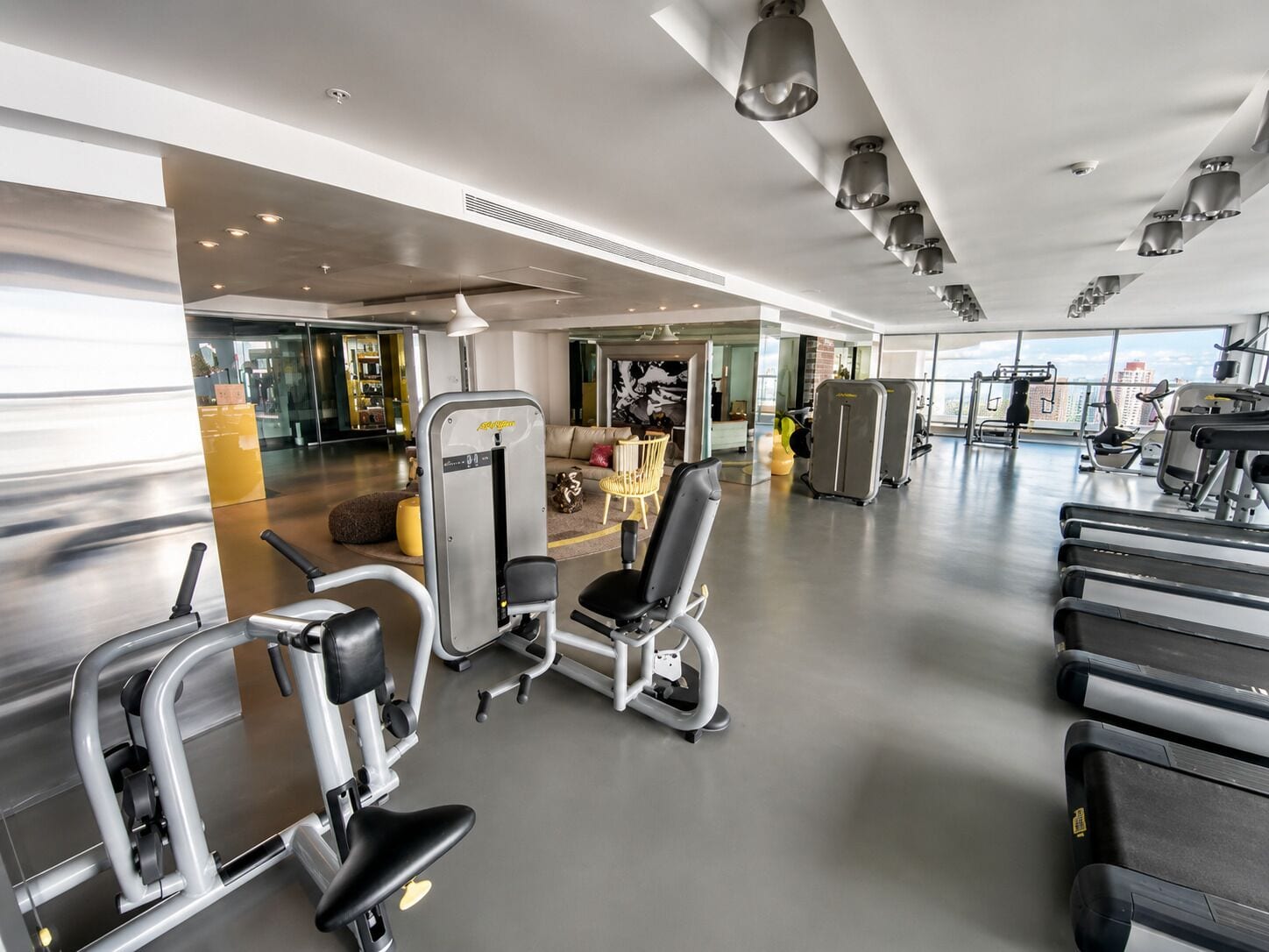 Une salle de sport moderne équipée de divers appareils d’exercice, dotée d’un espace détente et de grandes fenêtres offrant une vue sur la ville.