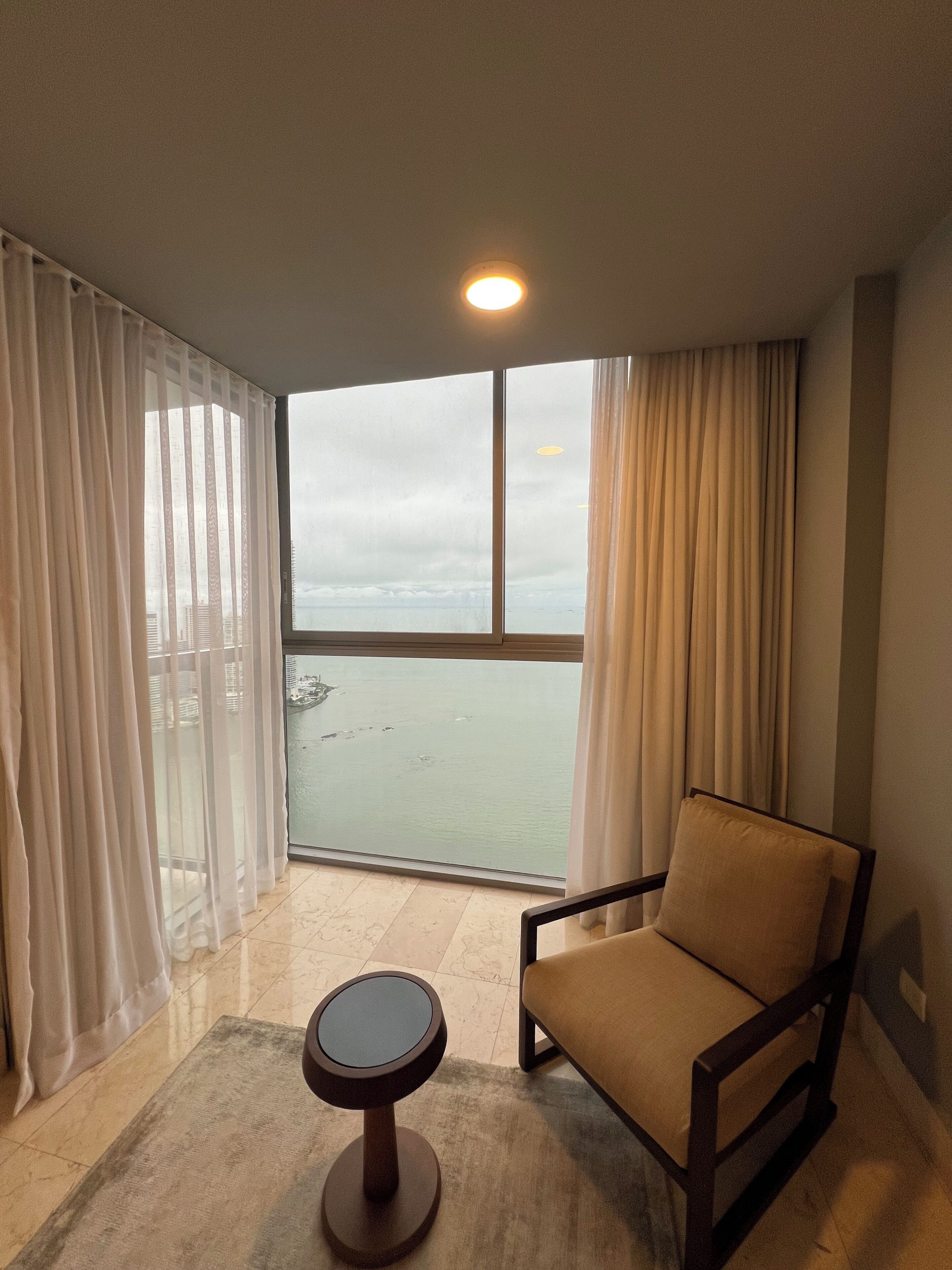 A room with a view of the sea, a chair, a small table, and sheer curtains.