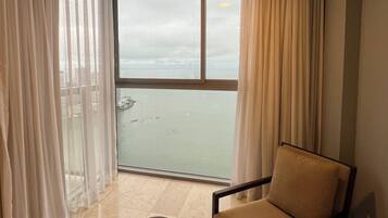 A room with a view of the sea, a chair, a small table, and sheer curtains.