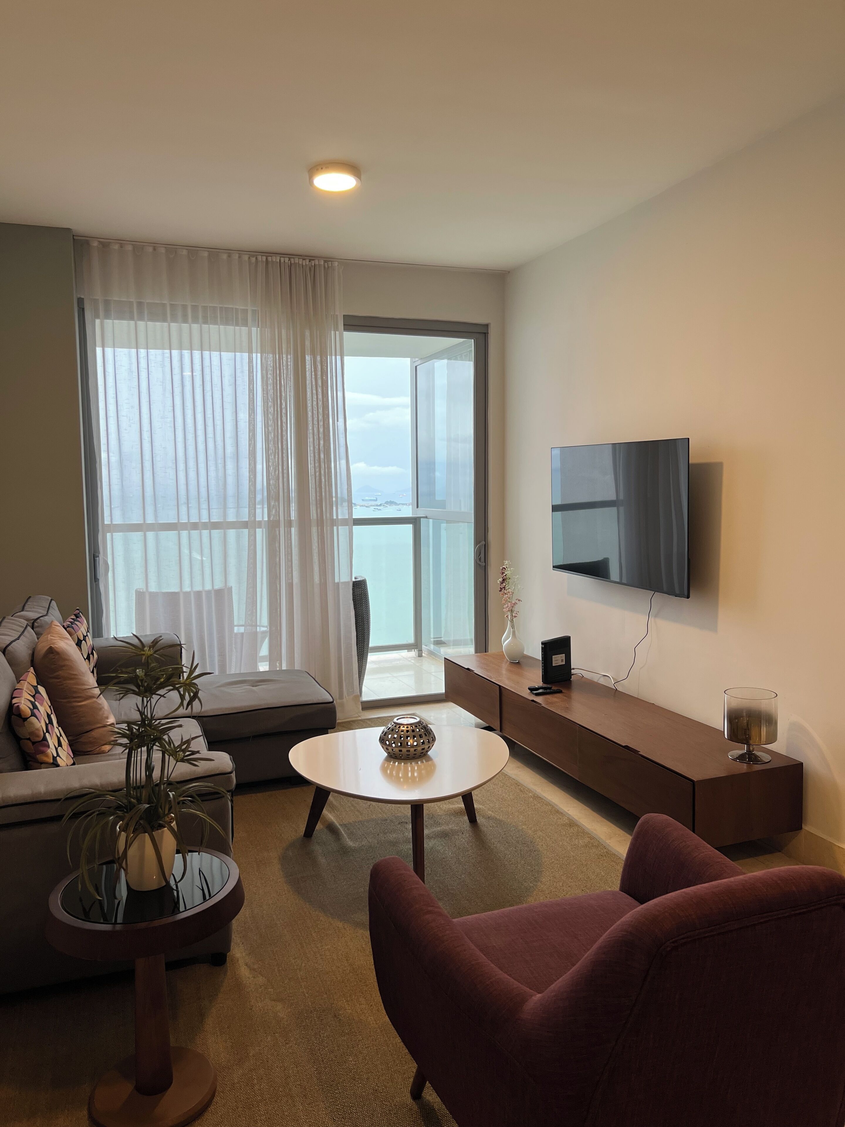 Ein modernes Wohnzimmer mit einer Couch, einem runden Couchtisch, einem roten Sessel, einem an der Wand montierten Fernseher und einem großen Fenster mit Vorhängen.