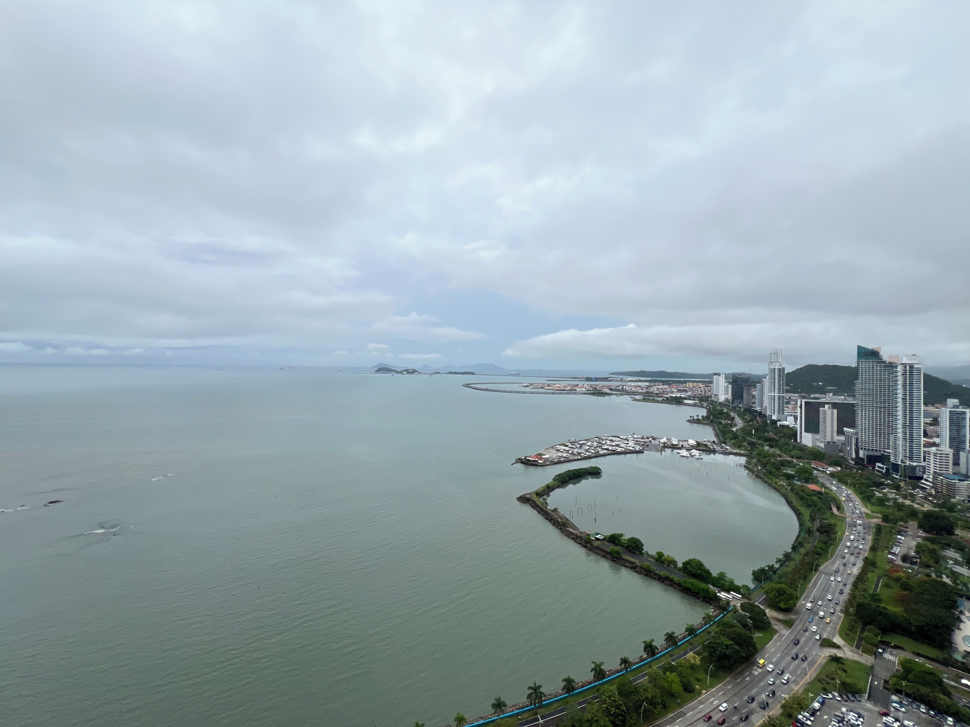 A coastal cityscape with a marina, high-rise buildings, and a curved road along the waterfront.