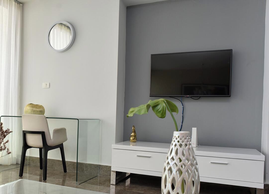 A modern living room with a flat-screen TV mounted on the wall, a glass coffee table, a white cabinet, and a decorative vase with a plant.