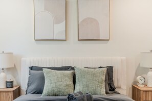 Une chambre à coucher avec un lit, des oreillers, une table de chevet, une lampe et deux œuvres d’art encadrées accrochées au mur.
