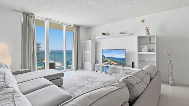 A modern living room with a large flat-screen TV, a sofa, and a balcony with a view of the sea. - New New  Tides 12E BeachView (Hollywood)