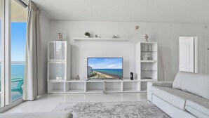 A modern living room with a large flat-screen TV, white furniture, and a view of the ocean. - New New  Tides 12E BeachView (Hollywood)