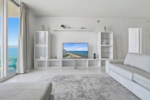 A modern living room with a large flat-screen TV, white furniture, and a view of the ocean. - New New  Tides 12E BeachView (Hollywood)