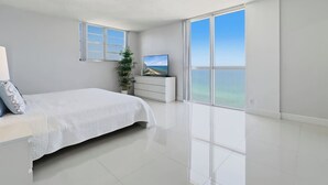 A modern bedroom with a bed, a TV, a plant, and a large window with a view of the ocean. - New New  Tides 12E BeachView (Hollywood)