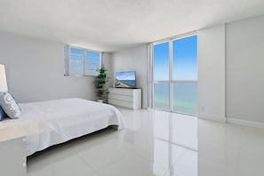 A modern bedroom with a bed, a TV, a plant, and a large window with a view of the ocean. - New New  Tides 12E BeachView (Hollywood)