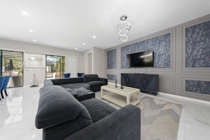 Ein modernes Wohnzimmer mit einem Ecksofa, einem Couchtisch, einem flachbildfernseher an der Wand und einem großen Fenster mit Blick ins Freie.