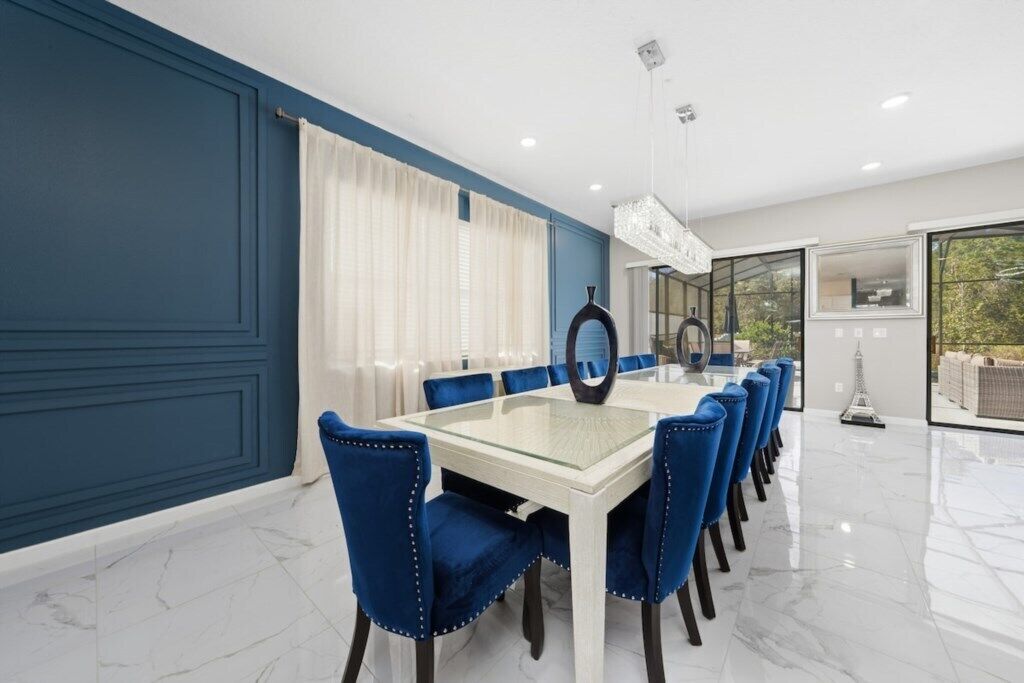 A dining area with a glass table and blue upholstered chairs, a chandelier, and a view through large windows.