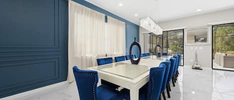 A dining area with a glass table and blue upholstered chairs, a chandelier, and a view through large windows.