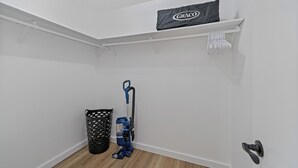 Una esquina de una habitación con una pared blanca, un bote de basura negro, una aspiradora azul y un protector de colchón Graco en un estante.