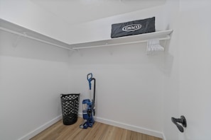 Una esquina de una habitación con una pared blanca, un bote de basura negro, una aspiradora azul y un protector de colchón Graco en un estante.