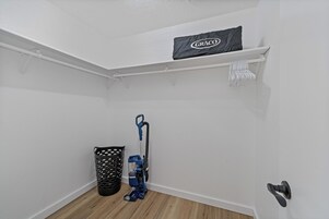 Una esquina de una habitación con una pared blanca, un bote de basura negro, una aspiradora azul y un protector de colchón Graco en un estante.