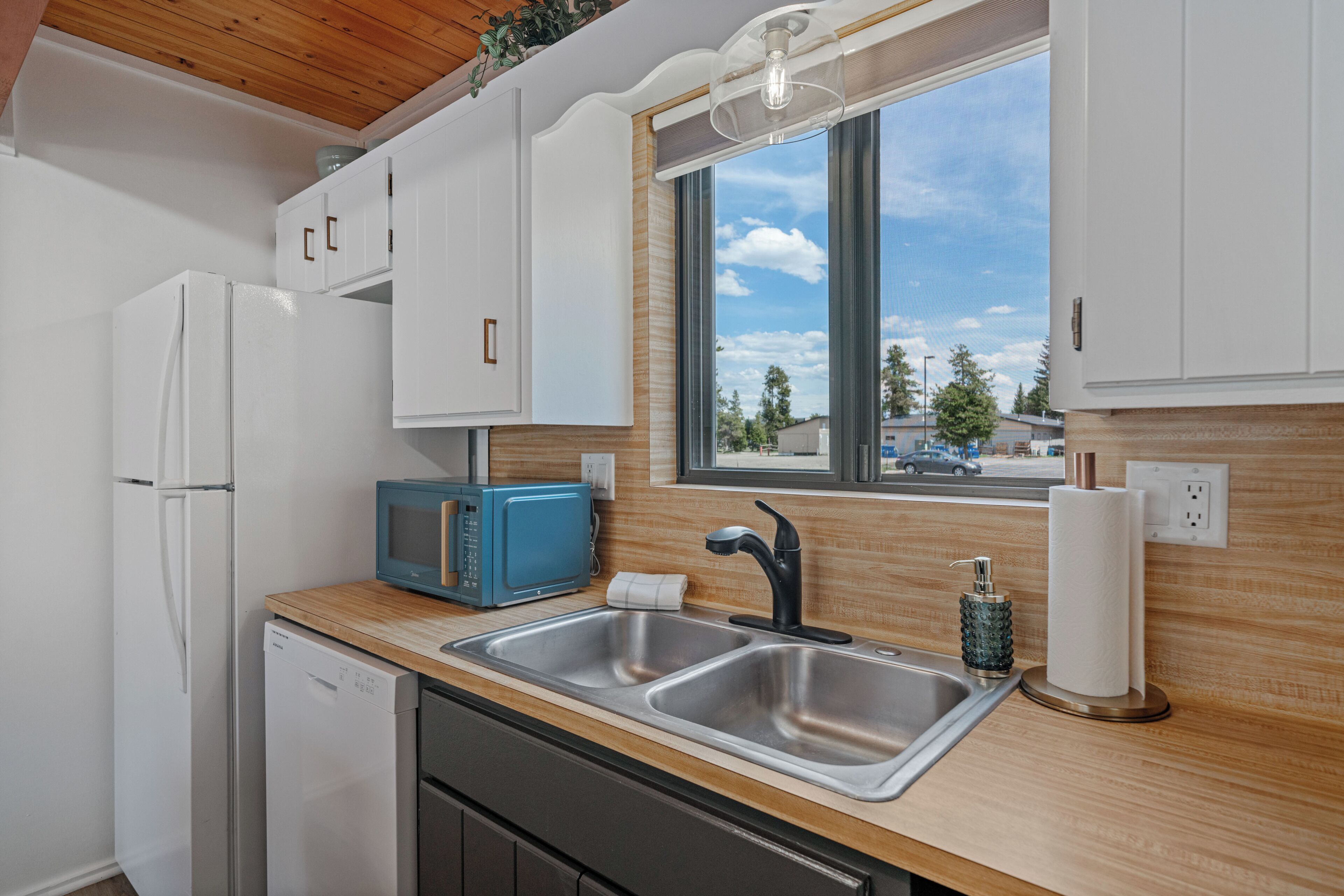 A modern kitchen with white cabinets, a blue microwave, stainless steel sinks, and a window with a view of the street.