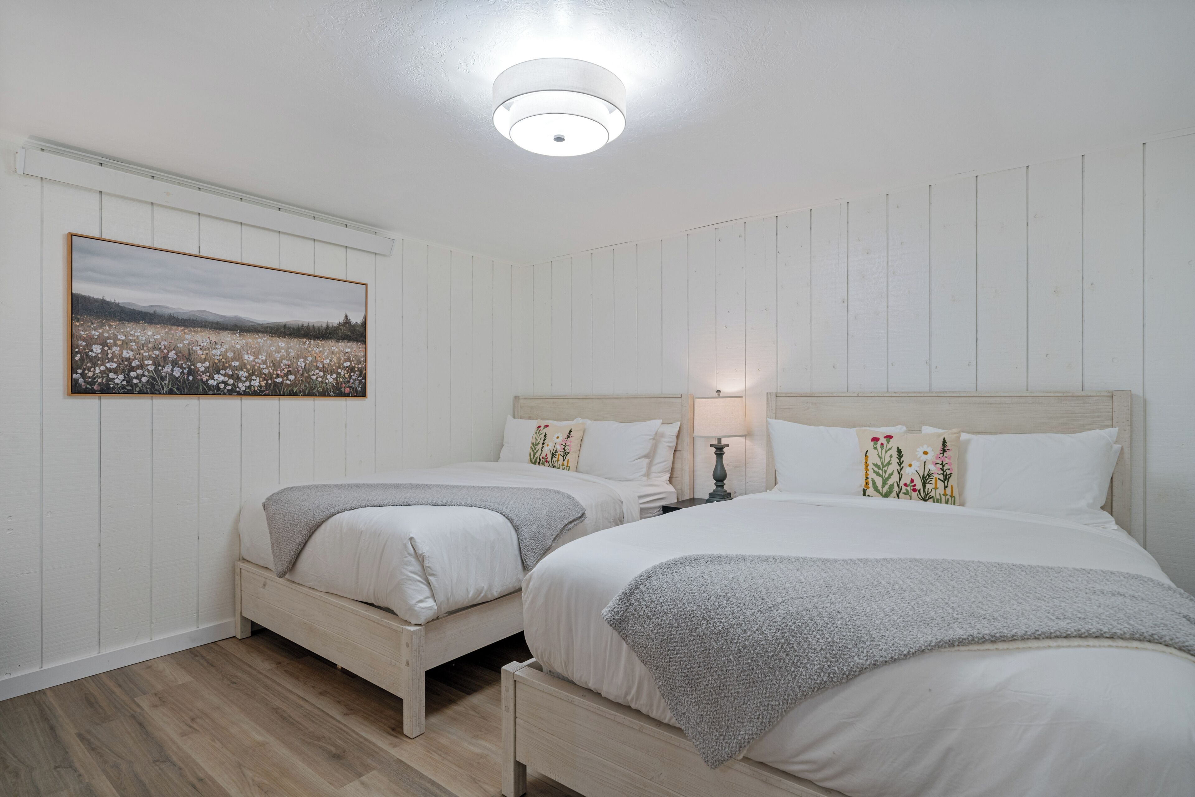Une chambre avec deux lits, un plancher en bois, un luminaire au plafond et un tableau accroché au mur.