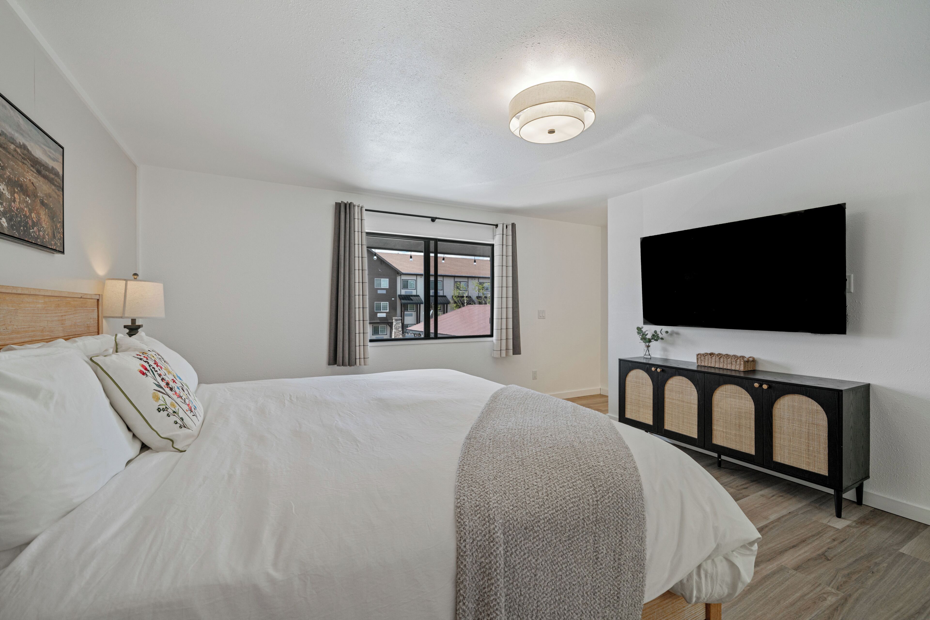 Un dormitorio con una cama, televisión instalada en la pared, una ventana con cortinas y una mesita de noche.