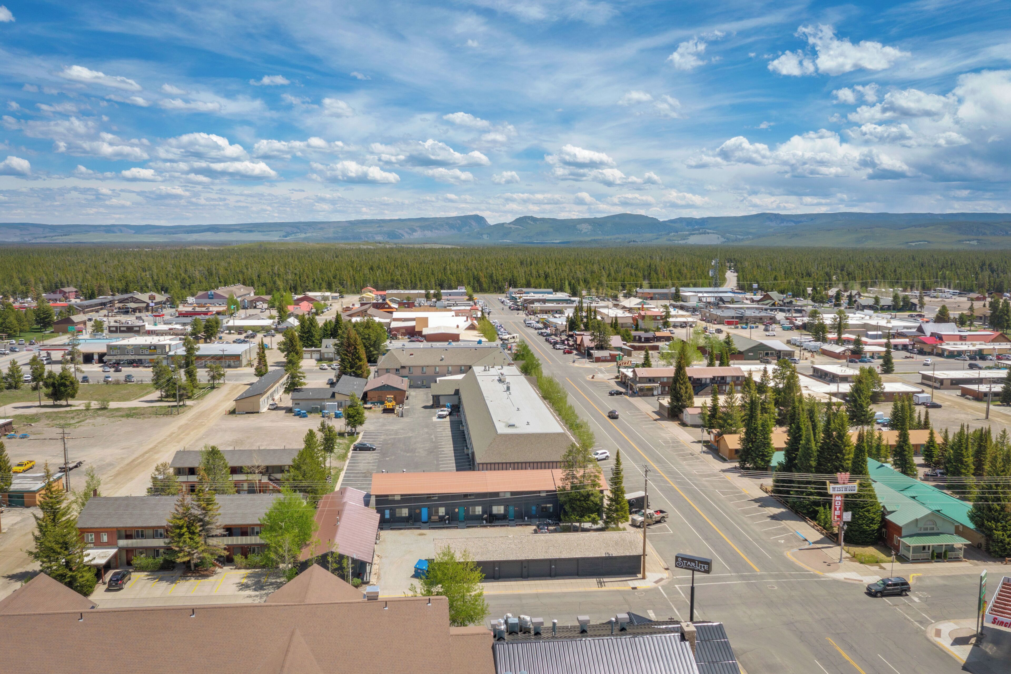 An aerial view of a town with multiple buildings, roads, and a forested area in the background.