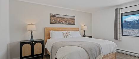 A bedroom with a wooden bed, two bedside tables with lamps, a wall-mounted painting, and a window with curtains.