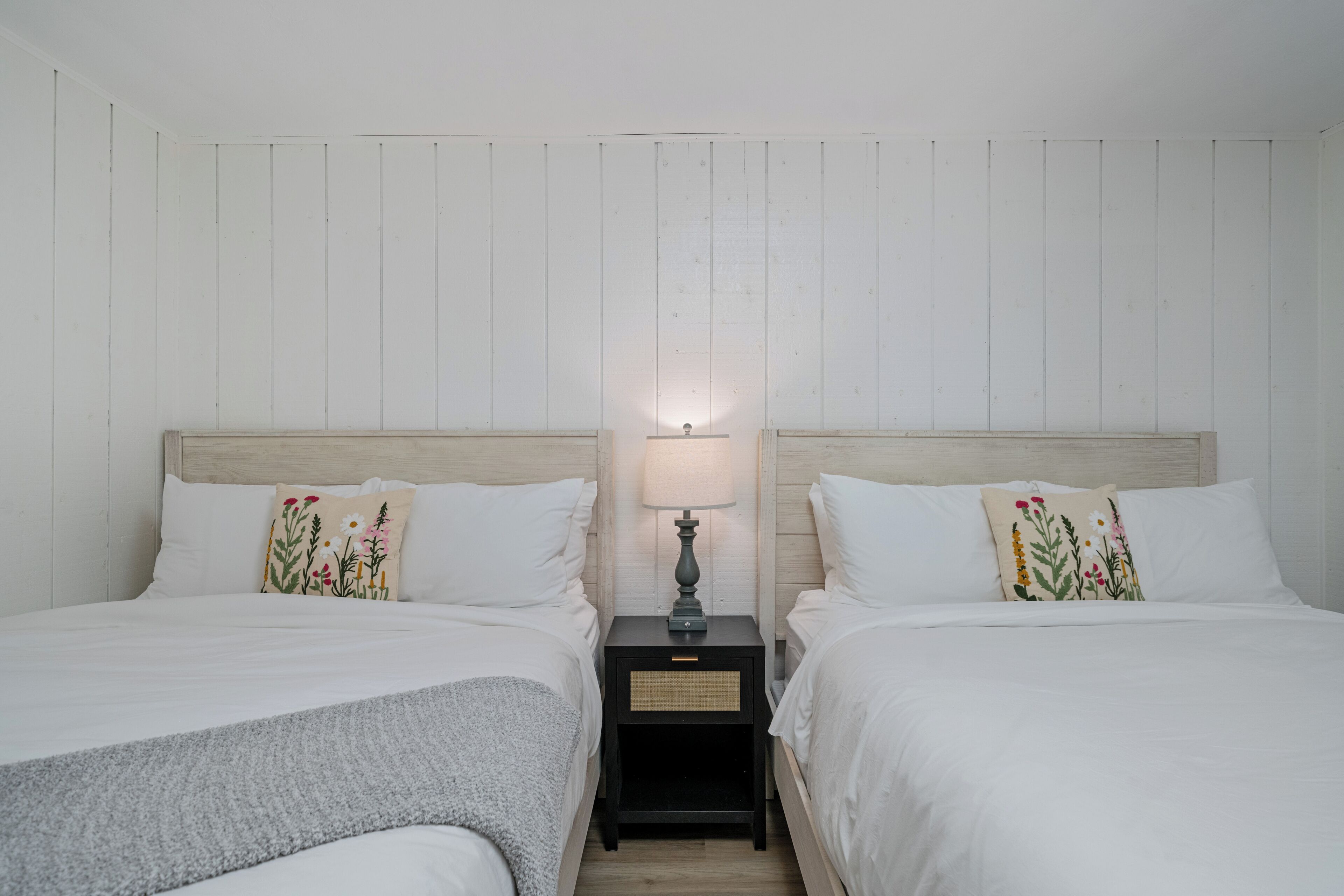 Una habitación con dos camas, una mesita de noche con lámpara y una pared con paneles verticales.