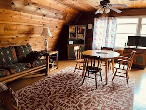 A cozy cabin interior with a wooden ceiling, a dining table with chairs, a sofa, a lamp, and a television.
