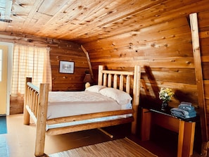 A wooden cabin room with a bed, a nightstand, and a window with curtains.