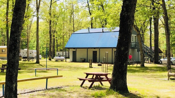 Um camping com mesas de piquenique, uma cabana com telhado azul e árvores ao redor.