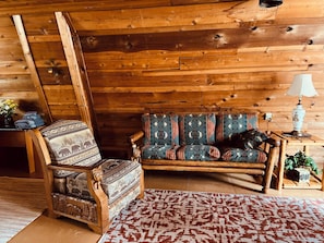 Interior de una cabaña de madera con una silla de madera, un sofá estampado, una lámpara y una alfombra.