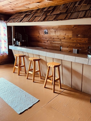 A wooden interior with a bar area, stools, and a patterned rug.