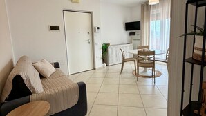 A modern living room with a sofa, a dining table with chairs, a TV mounted on the wall, and a kitchen area in the background. - Tourist Location Giancola between Seas and Mountains (Bellante Stazione)