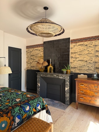 A room with a fireplace, a bed with a colorful bedspread, a wooden dresser, and a hanging light fixture.. Luxury B&B room in the center of Chinon