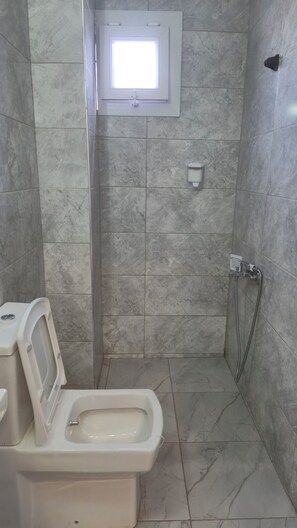 Uma casa de banho pequena, com azulejos, incluindo uma sanita, um chuveiro e uma janela.