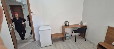 A small hotel room with a wooden desk, a compact refrigerator, a chair, a bed, and a trash bin.