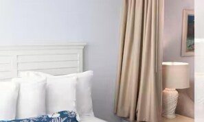 Habitació d'hotel amb un capçal de llit blanc, cortines beix, una làmpada blanca i un quadre al mur.