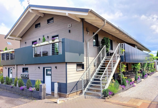 A modern two-story building with a visible staircase, a small balcony with plants, and a number '33' on the facade.