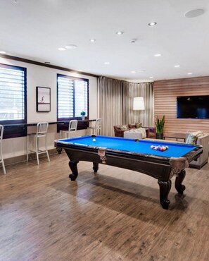 Una mesa de billar con superficie de fieltro azul, rodeada de piso de madera, un sofá y un televisor fijado a un panel de pared de madera.
