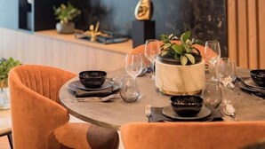 Een eethoek met een ronde tafel, oranje stoelen, een aan de muur bevestigde televisie en een moderne hanglamp.