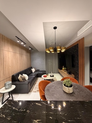 A modern living room with a sectional sofa, a round dining table with chairs, a small side table, and a ceiling light fixture.