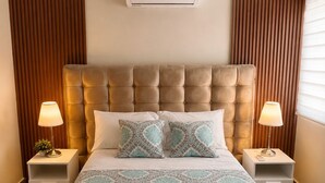 Quarto de hotel com cabeceira estofada, duas mesas de cabeceira com abajures, colcha estampada e ar condicionado fixado na parede.