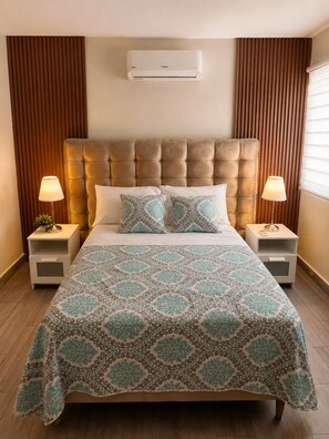A hotel room with a tufted headboard, two bedside tables with lamps, a patterned bedspread, and a wall-mounted air conditioner. - Warm and Cozy Apartment in Punta Cana (Punta Cana)