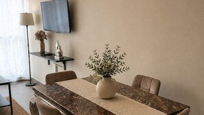 Uma sala de jantar com uma mesa de mármore, cadeiras acolchoadas, um vaso com folhagem, um abajur e uma televisão na parede.