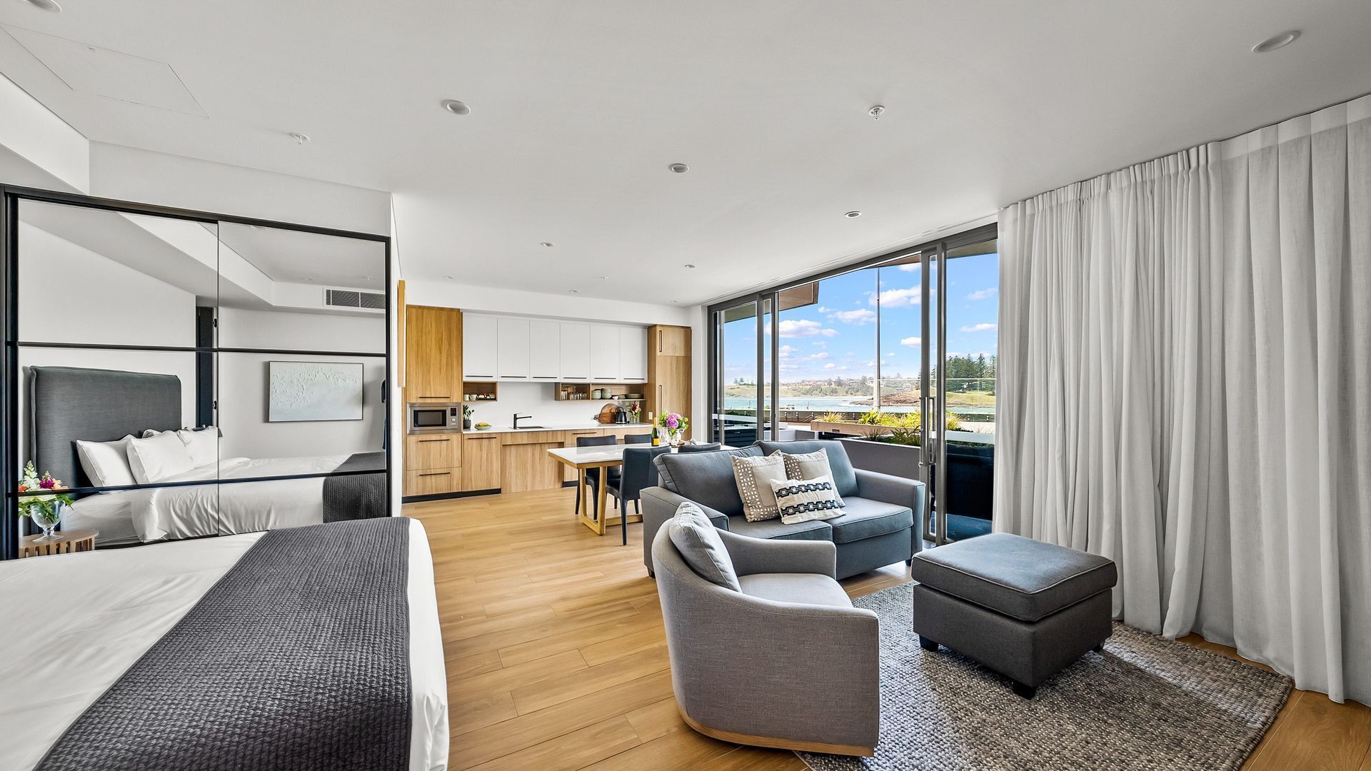 A modern hotel room with a bed, a sofa, a small table, a chair, and a view of the kitchen and outside.