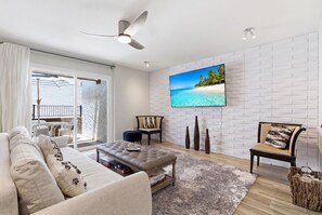 Ein Wohnzimmer mit weißer Ziegelwand, Flachbildfernseher, Deckenventilator, Sofa, Couchtisch und Blick auf einen Balkon.