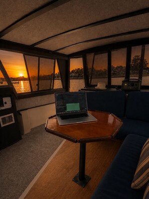 En bærbar computer på et bord i et rum med udsigt til solnedgang gennem store vinduer.