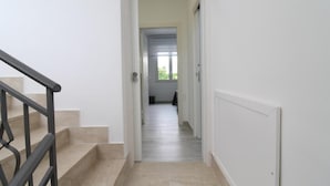 A hallway with a staircase leading to another room, white walls, and a door at the end.