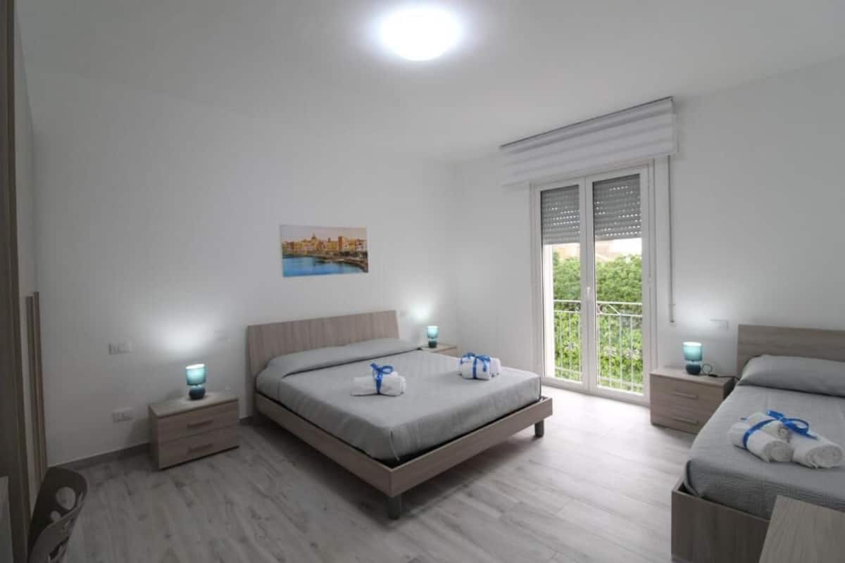 Un dormitorio con cama, mesitas de noche, ventana con vistas y un cuadro en la pared.