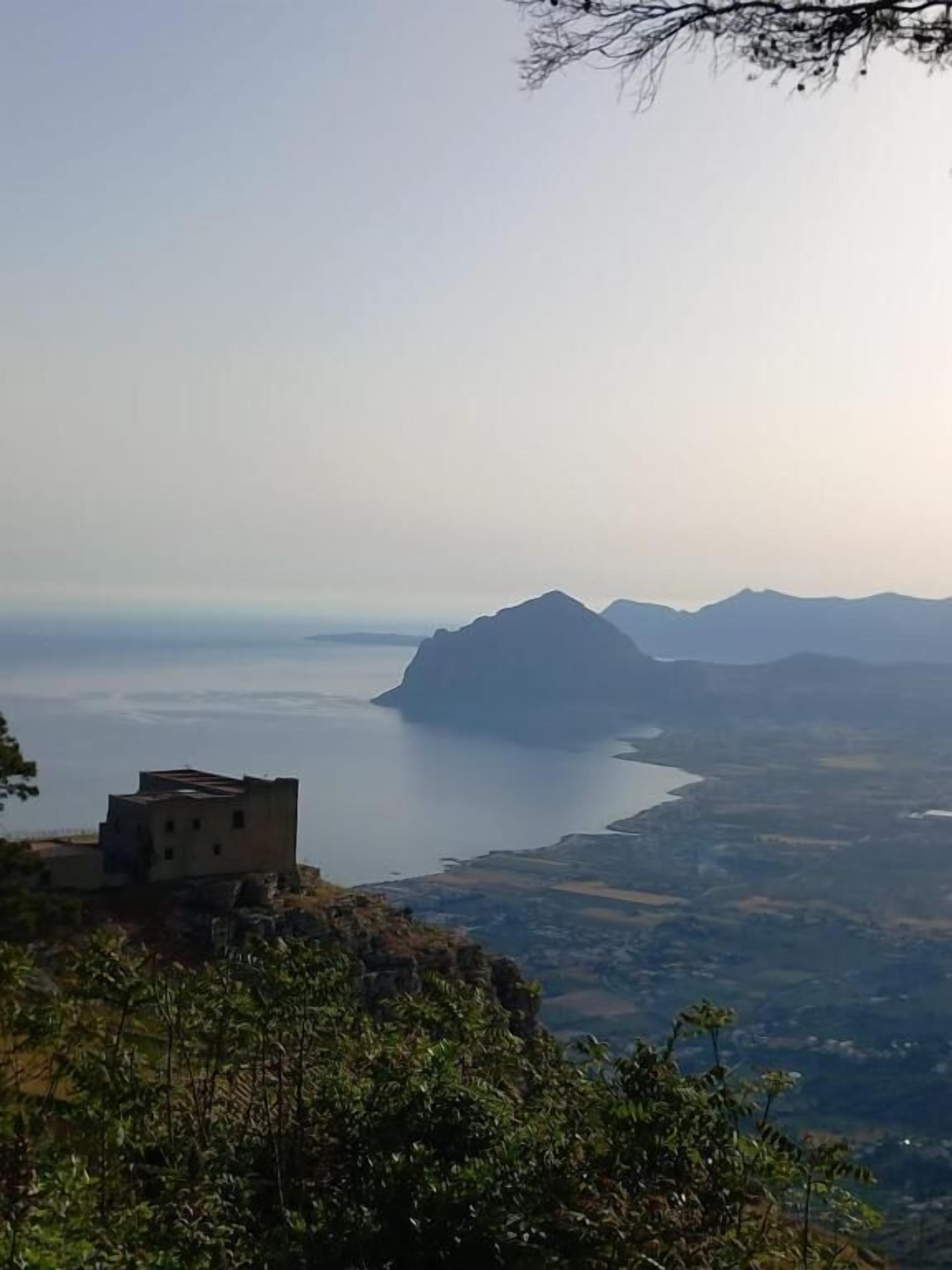 A coastal landscape with a view of the sea, mountains, and a building on a cliff.